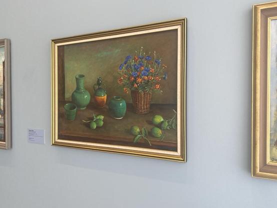 Still life by Margaret Olley. This one has orange and blue flowers in a wicker vase/pot, green vases and green lemons (with only a hint of yellow).