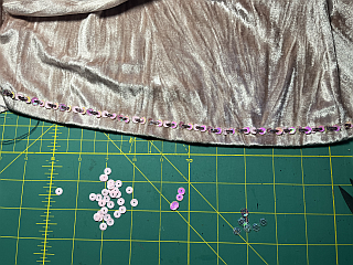 Nahaufnahme: Auf einer grünen Schneidematte mit gelbem Raster liegt in der oberen Hälfte altrosa-farbener Pannesamt. Am unteren Rand sind Perlen und Pailletten in verschiedenen Rosatönen aufgestickt.
Darunter liegen lose Pailletten in denselben Farben.