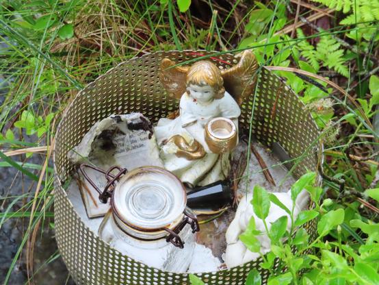 dans l'herbe au bord du ruisseau une sorte de panier métallique avec une statuette d'ange (blanc et doré) qui tient un pot, un bocal qui renferme une substance blanche, un morceau de papier imprimé abimé (brûlé ?) et d'autres matières blanches au fond. Très mystérieux !
In the grass by the stream, there is a kind of metal basket with a small statue of an angel (white and gold) holding a pot, a jar containing a white substance, a piece of damaged (burnt?) printed paper, and other white materials at the bottom. Very mysterious!