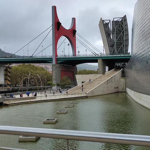 Blick auf das Guggenheim-Museum in Bilbao an einem bewölkten Tag. Rechts im Bild die geschwungenen, metallisch glänzenden Fassaden des Museums; links die markante rote La-Salve-Brücke mit ihrem Bogen über dem Nervión-Fluss. Auf dem Vorplatz steht die monumentale Bronzeskulptur Maman (1999) von Louise Bourgeois, eine übergrosse Spinne. Mehrere Personen spazieren entlang der Promenade und über die breiten Stufen am Wasserbecken.
