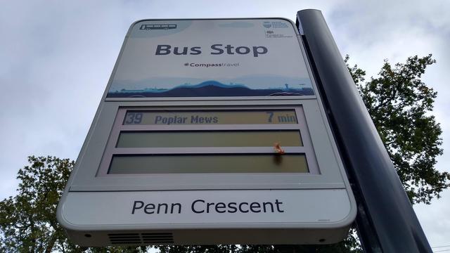 A view of a bus stop flag taken quite close and from below. At the top, it has the words "Bus Stop" over a strip depicting countryside and Gatwick Airport. At the bottom it says "Penn Crescent".

In the middle is a stack of 3 liquid crystal displays with the top one saying "39 Poplar Mews 7 min".
