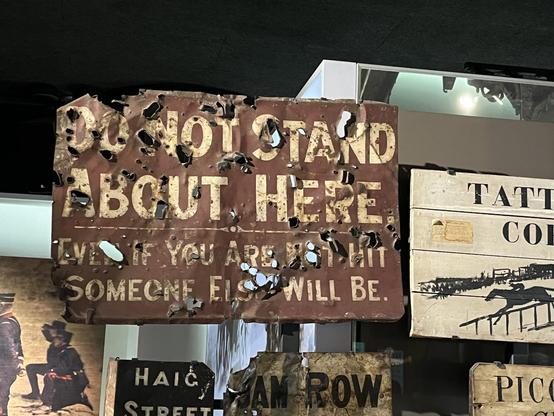 A heavily damaged sign, probably caused by shrapnel, saying: "Do not stand about here -- Even if you are not hit, someone else will be.