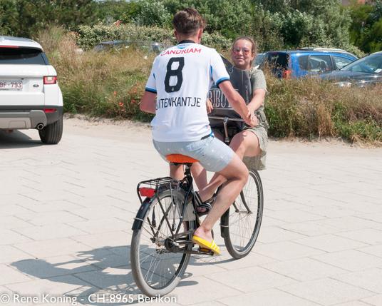Ein Fahrradfahrer fährt aus dem Bild. Er trägt ein T-Shirt mit der Rückennummer 8. In gegenübergesetzte Fahrtrichtung sitzt eine Junge Frau auf dem Schutzblech des Vorderrads, so fahren sie davon