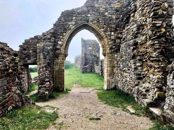 Ancient stone archway leads into the ruins of a medieval structure with weathered walls and partial openings visible. A grassy path runs through the foreground, bordered by walls consisting of various stones and bricks of different sizes and colors. The misty environment adds a serene atmosphere to the historical site.