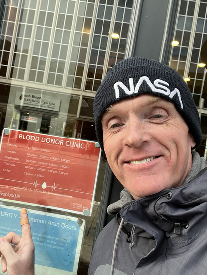 John A Mulhall in a grey jacket and NASA beanie hat pointing to an opening hours sign for blood donation clinic