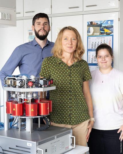 Foto: Ein technisches Gerät mit roten Komponenten auf einem Tisch im Labor, daneben stehen drei Menschen