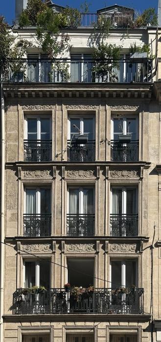 the side of an apartment building in the Parisian style, with three floors of floor-length windows with wrought-iron balconies, and above them, what appears to be an extension floor with a full-width balcony. There are plants on the balconies. The middle bottom window is open, exposing the darkness of the apartment.