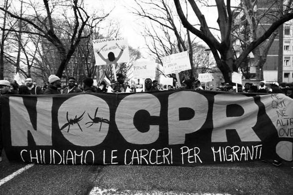 Un'immagine in bianco e nero di una manifestazione. Un grande striscione nero in primo piano recita "NOCPR CHIUDIAMO LE CARCERI PER MIGRANTI" (NO CPR CHIUDIAMO LE PRIGIONI PER MIGRANTI), con simboli di filo spinato all'interno della lettera "O". Dietro lo striscione, un gruppo di persone tiene cartelli, uno dei quali mostra una figura con le braccia alzate che spezza le catene e recita "VOGLIAMO PACE E LIBERTÀ". Altri cartelli visibili includono parole come "PERSONE" e "ITALIA". Lo sfondo mostra alberi spogli e edifici.
PH: Luca Greco