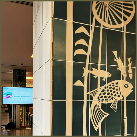 Foto: gekachelte Säule mit Fischen und Mustern, dahinter Screen mit Tagungslogo