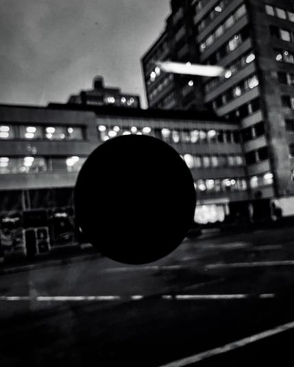 A blurred black circle in the foreground with illuminated buildings behind, captured in moody black and white at dusk.
