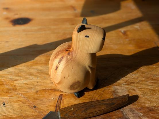 Woodcarved sculpture of a really round capybara on a workbench, next to a carving knife. Seen from its left side