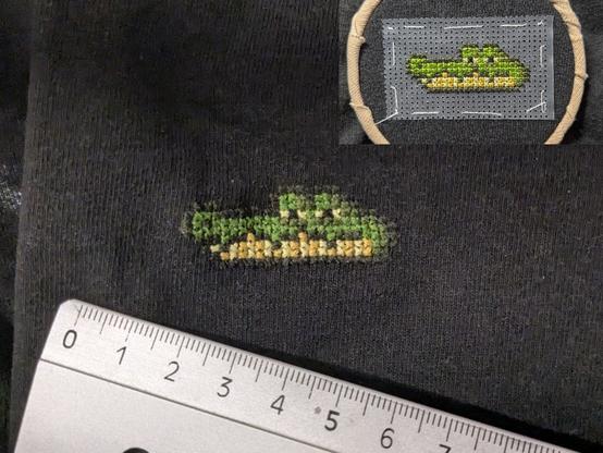 A toony crocodilian head hand embroidered on a black Tshirt fabric. It's done with cross stitch and looks blocky kinda like pixel art. There is a smaller picture top right with the embroidery still in the embroidery loop and with the cross stitch support layer (looks like a plastic sheet with a regular hole grid pattern).