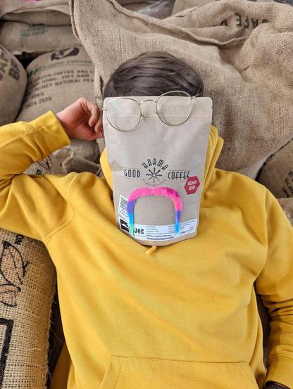 ein Bild mit dem Oberkörper einer Person die auf Kaffeesäcken liegt und eine Hand an den Kopf führt. über dem Gesicht liegt eine 250g Tüte Kaffee mit dem good karma logo, auf dem ein Regenbogenfarbiger Schnauzbart aufgeklebt ist. die Brille der Person ist etwas scheps auf die Kaffeetüte gesetzt.