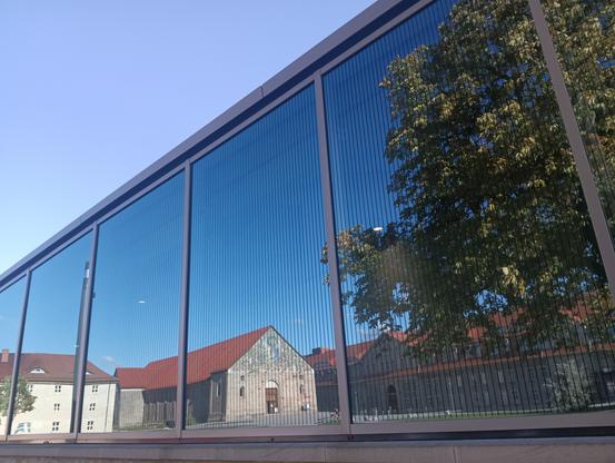 Foto der Fensterscheibe des Besucherzentrums auf dem Petersberg in Erfurt. 

In der Fensterscheibe, (Nordostseite) mit Sonnenschutzstreifen, spiegelt sich auf der linken Seite das Museum "Paradiesgärten – Gartenparadiese", ein Teil des Thüringischen  Landesamtes und ein Teil des Museums 
"Erlebnisportal". Auf der rechten Seite ist ein grün belaubter Baum. 
Das Foto wurde schräg aufgenommen, die untere Gebäudekante verläuft parallel zum Bildrand.