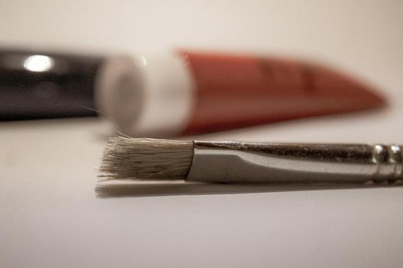Das Bild zeigt einen liegenden Pinsel in Nahaufnahme, dessen Borsten im Vordergrund klar erkennbar sind. Dahinter verschwimmen unscharf zwei Farbtuben, eine in Schwarz und eine in Rot. Die Szene vermittelt einen ruhigen, kreativen Moment und deutet auf Malerei oder künstlerisches Arbeiten hin.