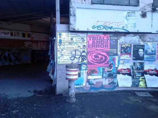 Foto vom Bauzaun der Scheinbaustelle der geplanten Durchwegung durch die Ruine des Ihmezentrums von der Blumenauer Straße zur Ida-Arenhold-Brücke. An der Absperrung längst obsolete Wegweiser zu gar nicht mehr vorhandenen Einrichtungen der Stadt Hannover im Ihmezentrum, daneben ein Reklameplakat für eine Veranstaltung in frischen, leuchtendem Magenta. Das Motto der Veranstaltung lautet »Trial and Error«. Davor steht ein Hydrant im Design der frühen Siebziger Jahre, stark korrodiert.
