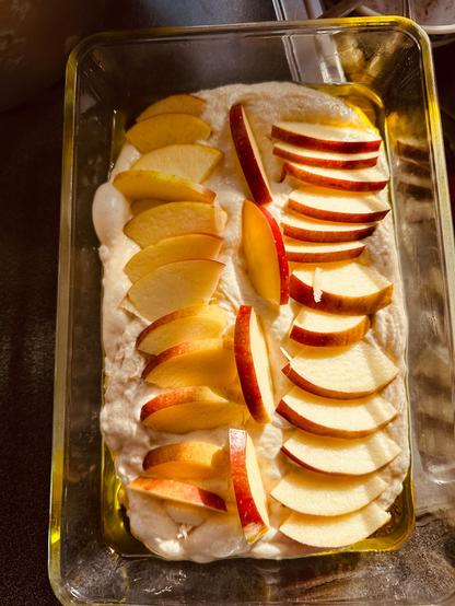 Sliced red and yellow apples are neatly arranged on a layer of dough in a rectangular baking dish.