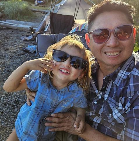 Smiling Asian man wearing sunglasses and a plaid shirt holding a smiling young child, also wearing sunglasses and a blue plaid shirt, at a campsite with a tent and beach in the background.