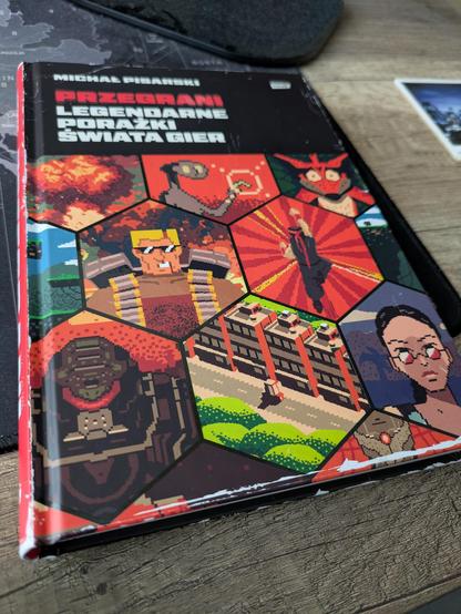 Zdjęcie książki "Przegrani. Legendarne porażki świata gier" autorstwa Michała Pisarskiego, wydanej przez Wydawnictwo Znak. Na brzegach kolorowej okładki widać mocne i wyraźne białe obdrapaną i rysy, ale one są nadrukowane.