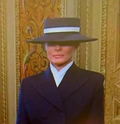 The First Lady wearing a black suit and black flat brimmed hat that covers her eyes. Her expression is, forgive me, inscrutable.
