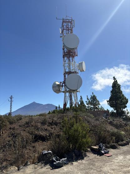 A telecommunications tower with large satellite dishes stands on rocky terrain. In the background, a Teide mountain is visible under a clear blue sky. There are some bags and amateur radio equipment placed on the ground nearby, along with sparse vegetation.