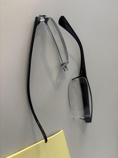 On a grey desk, with part of a pad of yellow sticky notes visible, are the broken pieces of a pair of plastic reading glasses. They broke in half at the bridge.