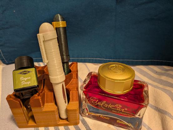 An inkstand carries two 3d printed pens and a bottle of Sugar Snap from Diamine. To the right a big bottle of Apricot Achat from Pelikan