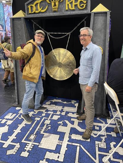 Vor einem großen Gong: Ich mit Klöppel eines Gongs, ausholend, auf der anderen Seite des Gongs ein ebenso wie ich bebrillter, aber glattrasierter Mann mit grauem Haar und Seitenscheitel, der grinst (Joseph Goodman von Goodman Games).