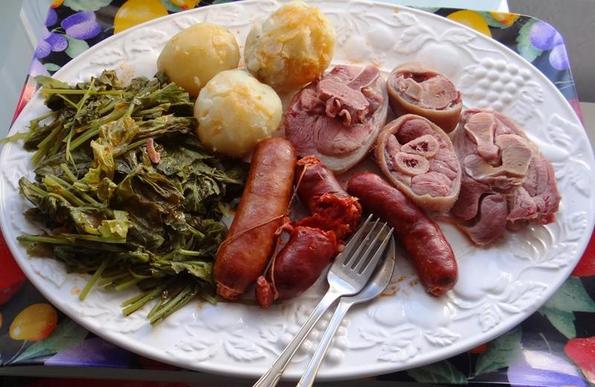 Un plato blanco rebosante de cocido gallego. En el plato se ven grelos, patatas cocidas (cachelos), varios tipos de chorizo y lacón cortado en rodajas, listo para comer.