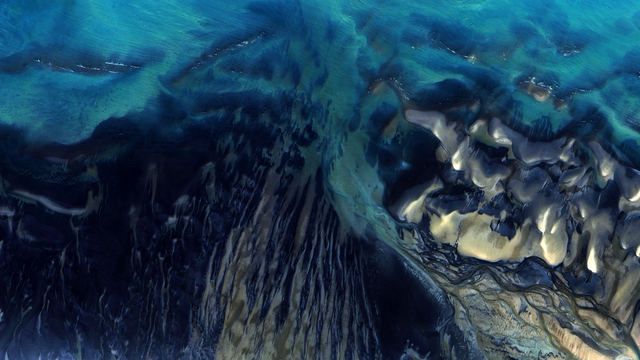 A satellite picture of the seawater on a coastline, with dark blue and teal tones interweaving in abstract patterns.