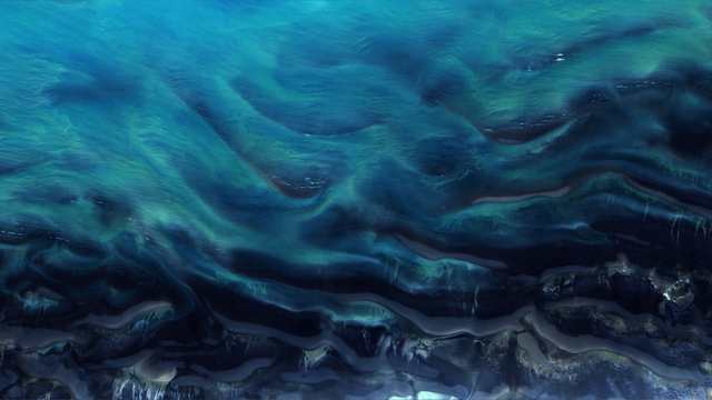 A satellite picture of the seawater on a coastline, with dark blue and teal tones interweaving in abstract patterns.