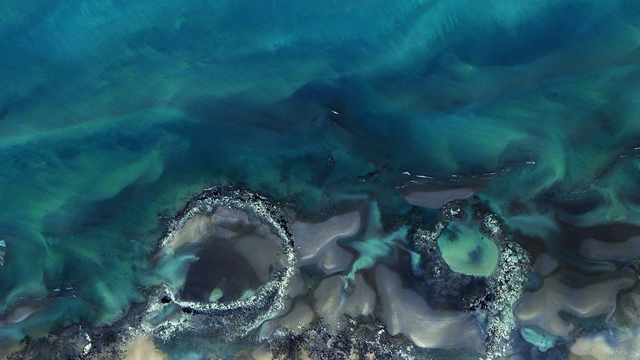 A satellite picture of the seawater on a coastline, with dark blue and teal tones interweaving in abstract patterns.