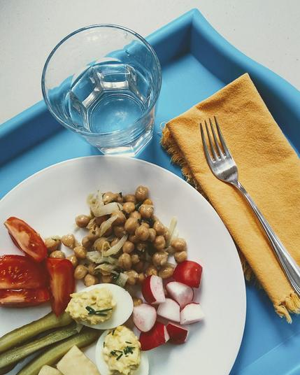 A blue tray set with a water glass, orange napkin, fork, and plate of food with chick peas, radishes, devilled eggs, pickles, and tomatoes.