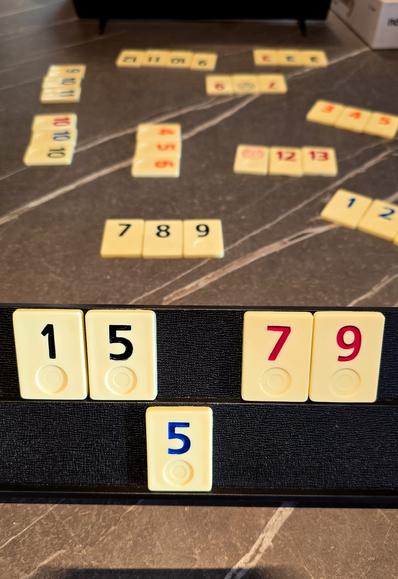 Ein Spielbrett mit Rummikub-Steinen auf einem Tisch. Im Vordergrund steht eine schwarze Ablage mit den Spielsteinen 1, 5, 7 und 9 sowie einer blauen 5 darunter. Im Hintergrund liegen weitere Zahlenkombinationen auf dem Tisch verteilt.