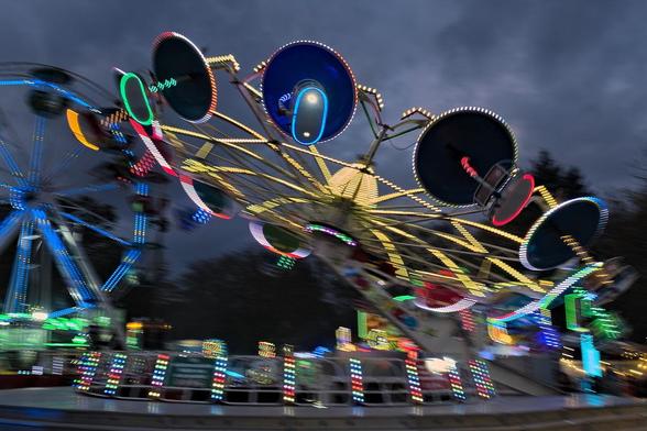 Ein Karussell, das leicht gekippt vor dem sehr grauen verhangenen Himmel dreht. Die Gondeln sind sehr klein, für zwei Personen, und darüber mittels gebogenenr Stange ein relativ großes Dach, wie ein Schirm. Die Gondeln sind alle in unterschiedlichen bunt Farben beleuchtet. Durch die Bewegung sind alle unscharf ausser die Gondel in der Bildmitte, die unten zwei Leuchten hat, die in die Kamera leuchten.
Im Hintergrund ist ein Miniatur-Riesenrad