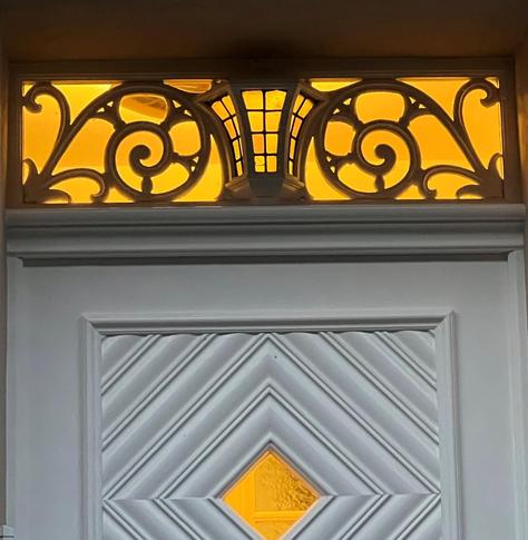 A decorative door featuring a white panel design and an ornate transom above with yellow illumination. The transom includes intricate scrollwork and a central lantern-like motif.

Eine dekorative Tür mit einem weißen Plattendesign und einem verzierten Heck oben mit gelber Beleuchtung. Der Querrübergang umfasst komplizierte Schriftrollen und ein zentrales laternenartiges Motiv.
