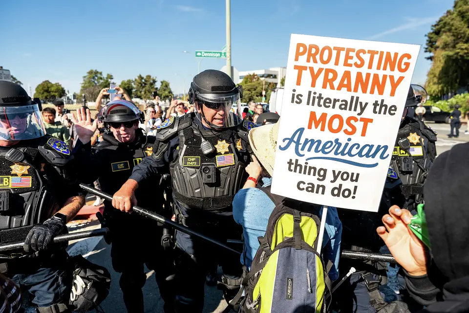 CHP officers in full riot gear pushing back a protesting crowd at the Alameda Airbase. One of the demonstrators is holding a sign, prominent in the photo, that reads: "Protesting Tyranny is literally the most American thing you can do!"