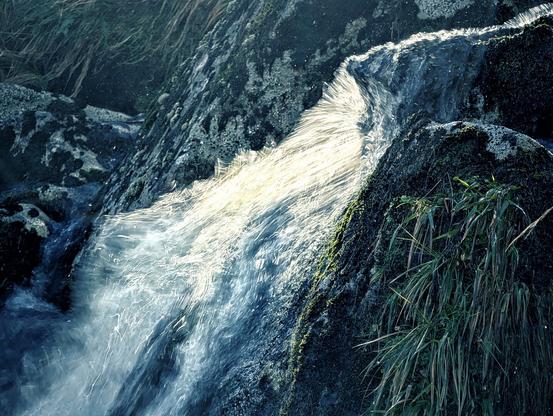 A close-up, low-angle photograph of water rushing over dark, moss-covered rocks. The water is captured with a slight motion blur, and a bright beam of sunlight is reflecting intensely off the centre of the cascade, giving the water an ethereal, almost luminous golden glow against the shadowed, deep blue and green tones of the surrounding rock and grass.