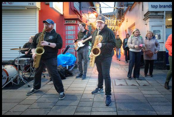 Two saxophone players from TBL8 Brass performing on Oliver Plunkett Street during Cork Jazz Festival with drummer and bassist visible in background, shoppers and spectators watching from the pedestrianised street at dusk with Christmas lights illuminating shop windows.