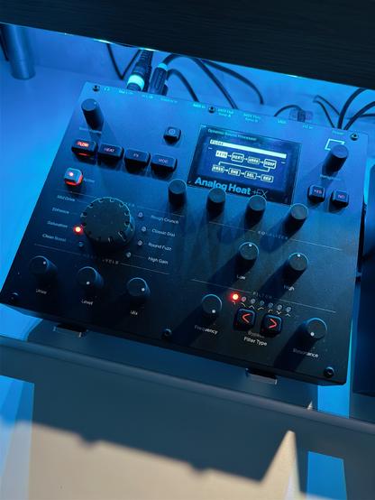Zařízení Analog Heat +FX, což je zvukový procesor. Zařízení má černou barvu a je osvětleno modrým světlem. Na horní části panelu se nachází displej zobrazující aktuální režim a parametry. Pod displejem jsou tlačítka pro výběr různých zvukových efektů, jako jsou FITS, HEAT, WARP a CORP. Dále jsou zde ovladače pro úpravu zvuku, včetně knoflíků pro úpravu hlasitosti vstupu a výstupu, ovládání efektů (FLOW, HEAT, FX, MOD), a různé typy filtrů a ekvalizérů. Vlevo se nachází velký otočný knoflík pro výběr úrovně a stylu efektu. Na pravé straně jsou tlačítka YES a NO pro navigaci a potvrzení voleb. Zařízení je připojeno několika kabely.