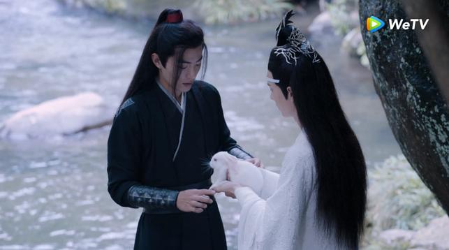 Wei Wuxian and Lan Wangji stand facing each other by a river in the Cloud Recesses. Wangji is carefully cradling a white rabbit in his hands, holding it out to Wei Wuxian. Wuxian has hesitantly brought his hands up beside Wangji's in preparation for receiving the rabbit.