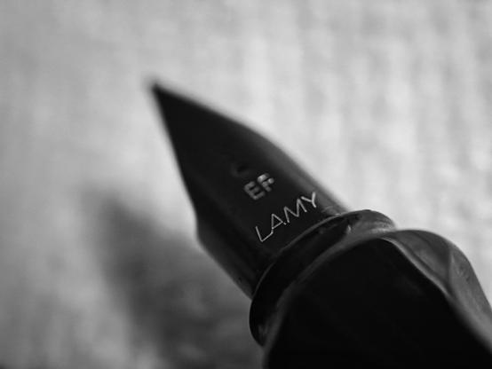 A close-up black-and-white photograph of a fountain pen’s pointed nib poised beside a blurred patch of ink on the surface of a white paper towel. The nib, bearing the fine text "EF LAMY," is in sharp focus on the right, while the ink spot and textured grid of the towel appear softly out of focus in the background. Light reflects off the shiny, dark metal of the nib, contrasting with the matte, fibrous texture of the absorbent towel below. The composition places the nib partially over the towel, suggesting the origin of the ink marks captured in the previous images.