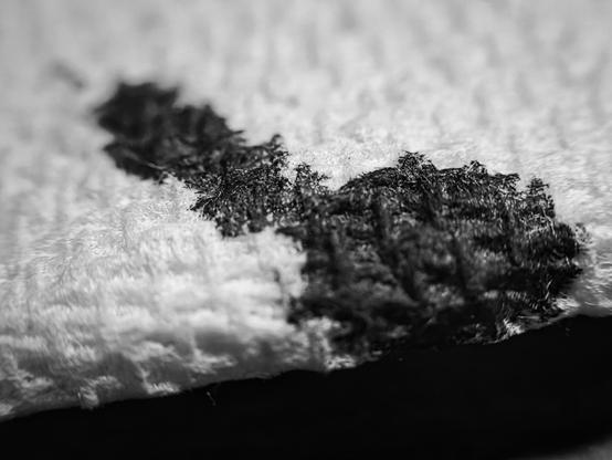 A black-and-white macro photograph showing a textured patch of dark ink absorbed into the surface of a white paper towel. The ink splotch appears irregular in shape, stretching diagonally from the upper left to the lower right, with frayed and fuzzy edges as the ink is wicked into the surrounding paper fibers. The extreme close-up captures the woven, dimpled structure of the towel’s fibers, some soaking up the ink and others remaining crisp and white. The lighting is soft and diffuse, highlighting the contrast between the deep black ink and the delicate grid-like pattern of the towel. The composition draws focus to the intersection of ink and fiber, revealing the micro-texture created by the ink’s interaction with the absorbent material.