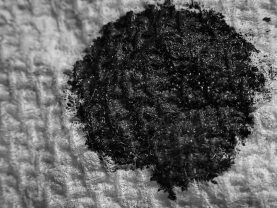 A monochrome macro shot of a circular black ink blot absorbed into a white paper towel, filling most of the frame with its irregular, fuzzy edges. Beneath the ink, the paper towel’s fine woven pattern remains visible, with the ink soaking into the fibers and softening the texture toward the edges. Light falls gently across the scene, revealing both the density of the central ink mass and the subtle gradient where the ink feathers into untouched towel. The background highlights the sculptural quality of the towel’s surface, with raised threads accentuated by the dramatic focus and side-lighting.