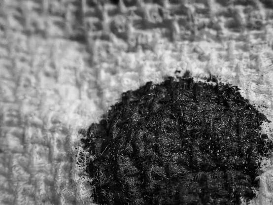 A black-and-white macro image of a bold, round splotch of black ink settled into the woven surface of a paper towel. The ink forms an almost perfect circle near the center, its edges feathered outward as it seeps into the towel’s porous texture. Fibers around the ink show fine, subtle detail, with the grid-like weave pattern providing a tactile backdrop. Soft, even lighting accentuates the difference between the saturated ink spot, dense and glossy, and the surrounding dry, matte paper towel. The edges of the ink mark create a delicate transition from black to gray, hinting at slight absorption and capillary spread within the towel.
