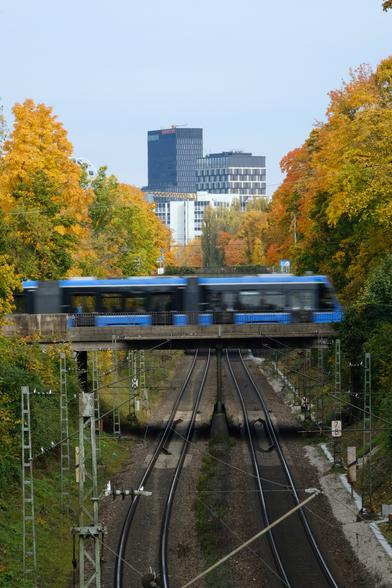 A view from a bridge over rails. The rails provide lines leading towards the center of the frame, to another bridge. This bridge in the center is framed in either side by beautiful yellow autumn trees. Across this bridge drives a blue tramway. The shutter speed is so long, that the motion blurs the driving tramway.