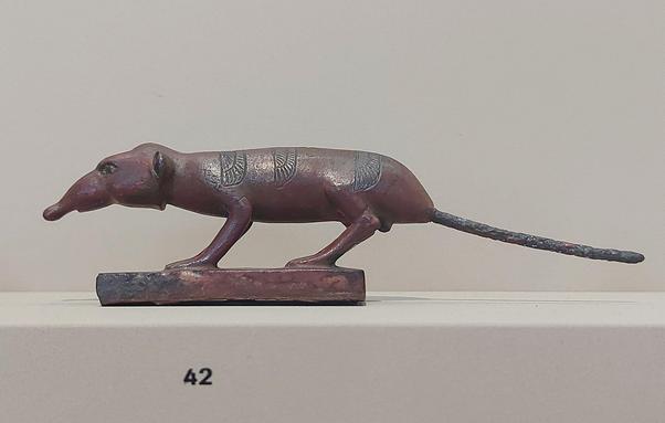 A small bronze statue of a shrew standing on a rectangular base. The figure is elongated with a pointed snout, slender legs, and a long tail, featuring engraved decorative patterns on its back. Displayed against a neutral background with the label number 42.
