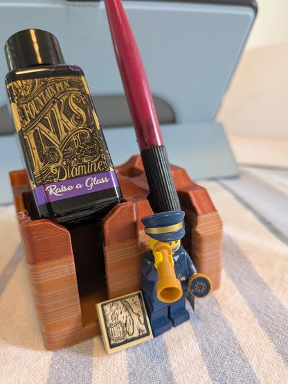 The pen and ink stand has a bottle of 'Raise A Glass' ink and a dip pen on it. In front a Lego minifig of a lighthouse captain holds a compass and telescope with a Lego element that could be a map on the tablecloth next to him.