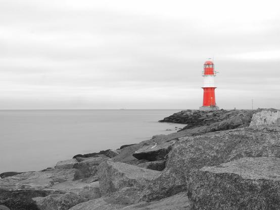 Ein Schwarzweißbild von einer Mole mit Molenfeuer. Das rot-weiße Molenfeuer ist das einzige Farbelement in dem Bild. Das Meer sieht durch Nutzung eines Graufilters komplett glatt aus. Der Himmel ist mit Wolken bedeckt.