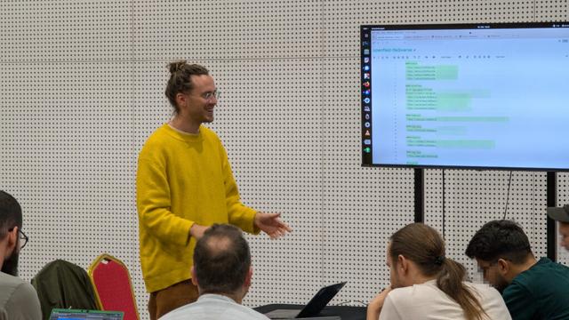 People sitting around a table. Some have notebooks to work on the workshops tasks. André is standing with a bright yellow pullover and explaining stuff. There is a big screen that shows some information about the workshop.